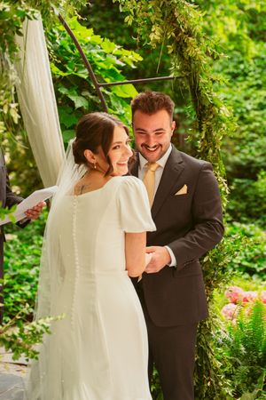 A wedding couple shares a joyful moment under an ivy-covered arch in a verdant garden setting.