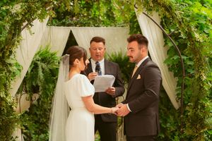 An outdoor wedding ceremony takes place under a white draped arch covered in green vines.