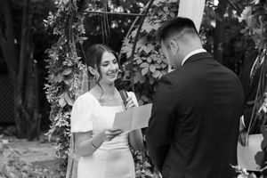 A couple exchanges vows during an outdoor wedding ceremony under a floral arch in black and white photography.