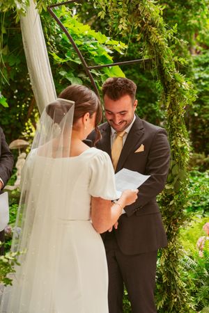 A couple reads their vows while standing beneath a decorated garden arch with white draping.