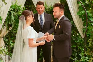 A couple exchanges rings during an outdoor wedding ceremony under a white draped arch with lush greenery.