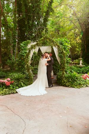 Newlyweds share intimate moment under floral-covered garden arch with pink roses and greenery.