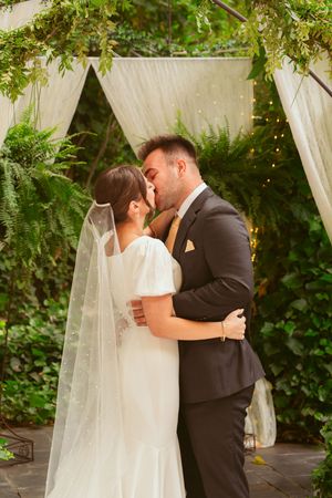 A romantic first kiss as newlyweds under a white draped archway surrounded by garden foliage.