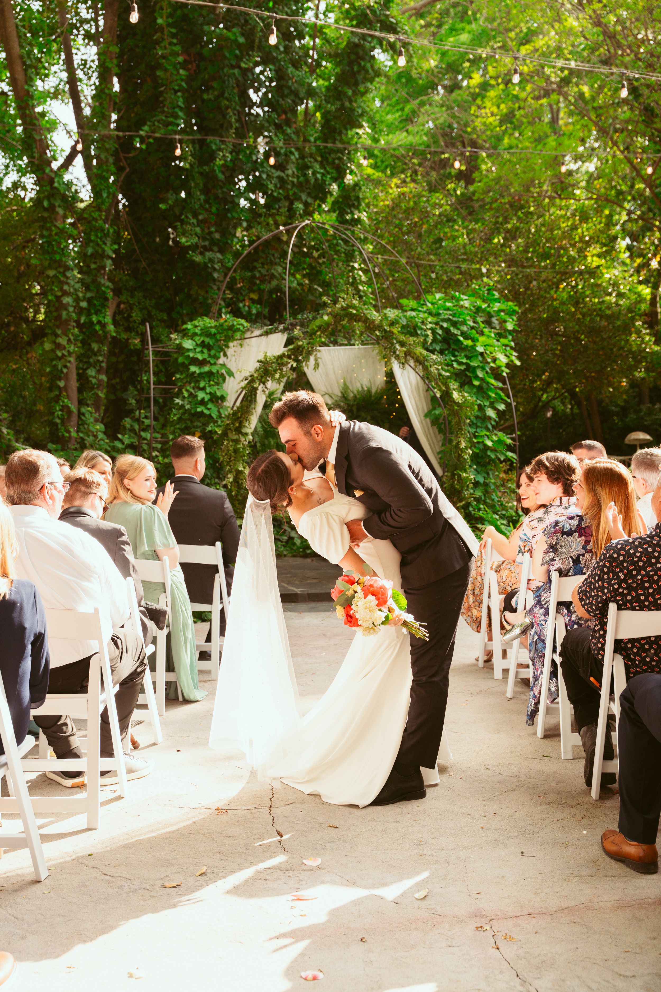 Wedding couple shares romantic kiss at outdoor ceremony with white chairs and green foliage backdrop.