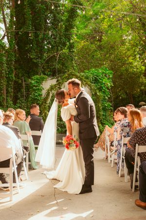 Newlyweds share their first kiss under a garden arch while wedding guests watch from white folding chairs.