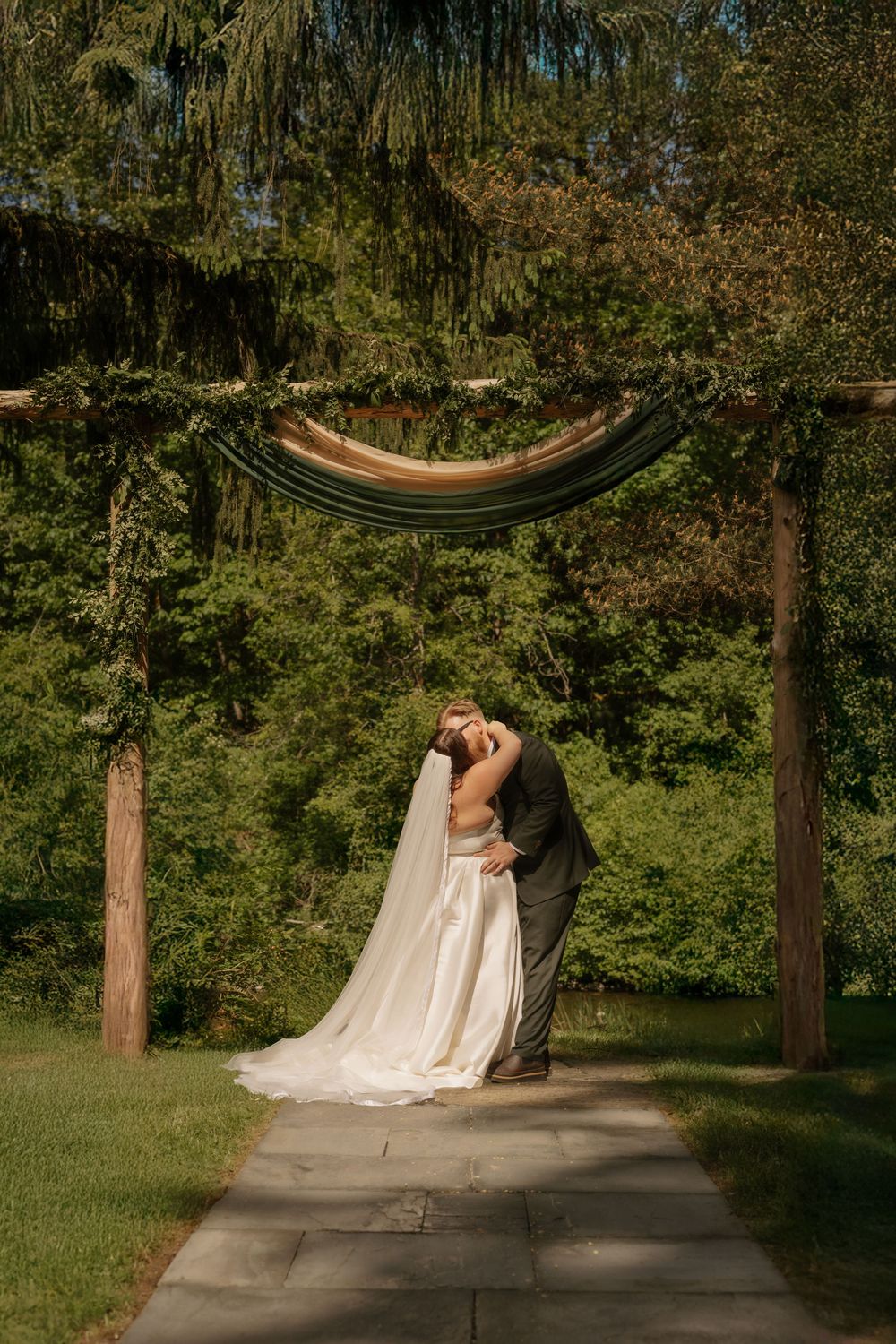 A couple shares a romantic moment under a curved wooden arch on a stone path surrounded by lush green gardens.