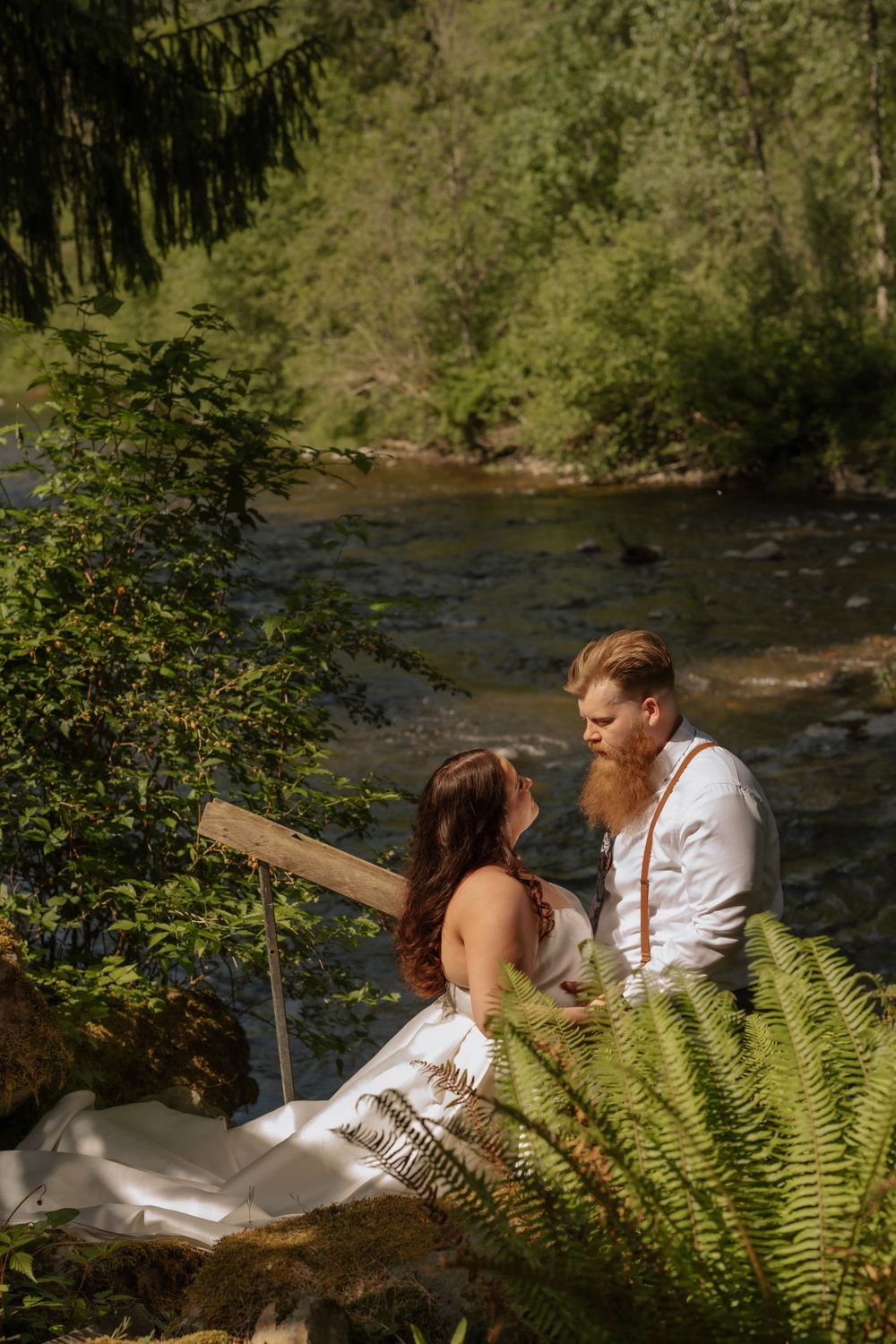 A romantic couple sits by a peaceful river surrounded by lush ferns and forest greenery on a sunny afternoon.