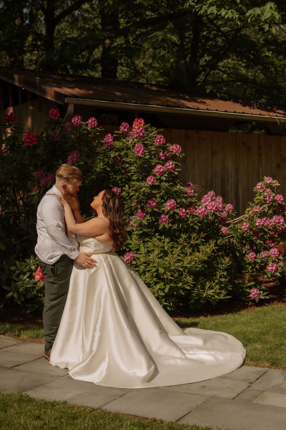 A couple shares an intimate moment on a stone patio surrounded by vibrant pink roses and a rustic wooden backdrop.