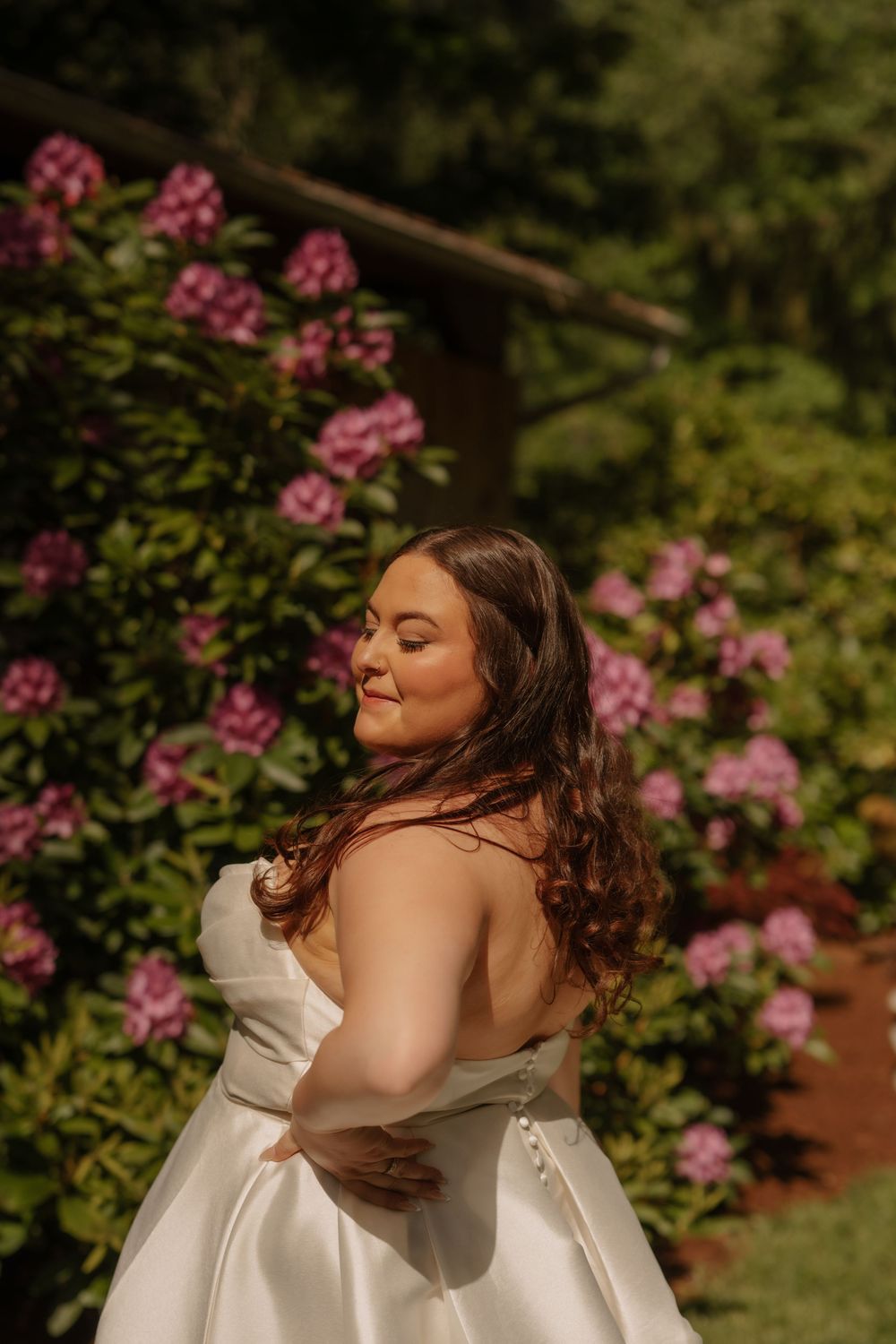 A bride in a white satin wedding dress poses in a garden with blooming pink flowers on a sunny day.