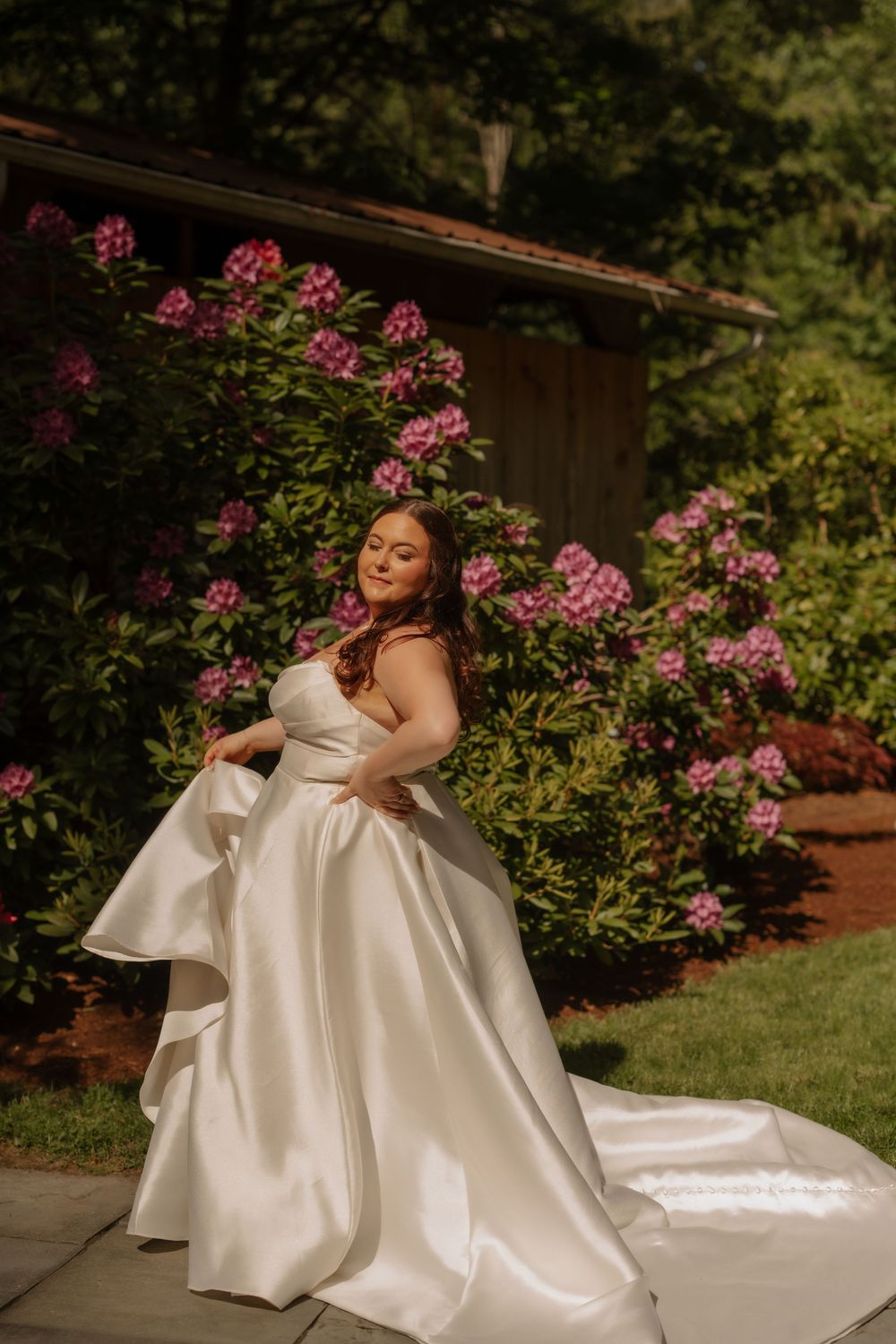 A bride in an elegant white satin ballgown poses outdoors next to blooming pink flowers and greenery.