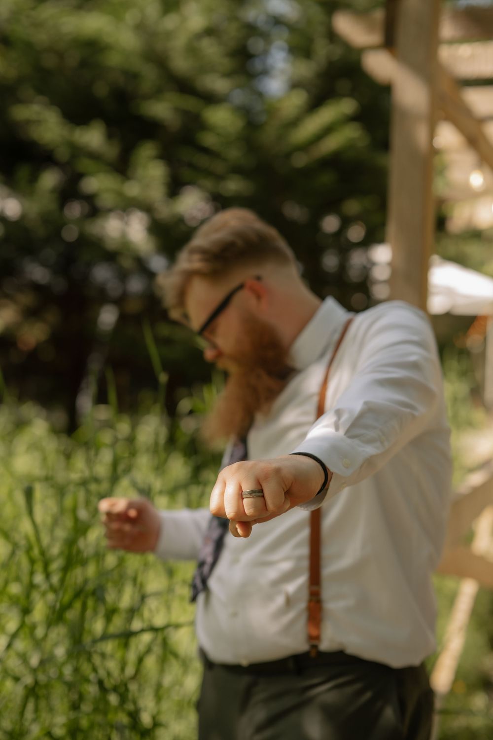 Man in white shirt with suspenders stands thoughtfully in a sunlit garden setting with blurred greenery behind him.