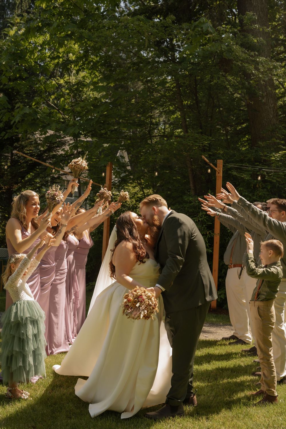Wedding party celebrates with confetti toss during outdoor ceremony under trees as couple shares romantic moment.