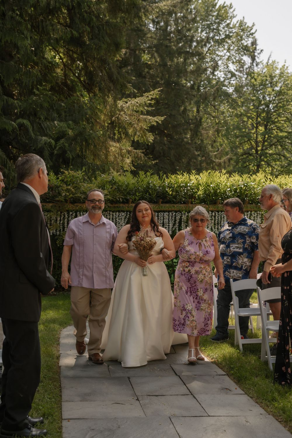 Wedding procession walks along stone pathway under trees as bride carries bouquet during outdoor summer ceremony.