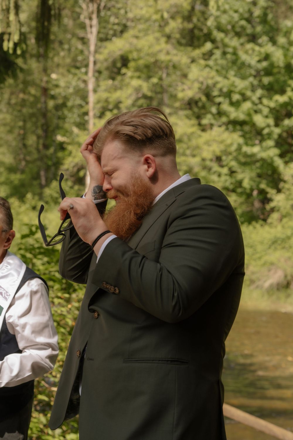 Emotional groom in dark suit wipes tear during outdoor wedding ceremony by a river in a forest setting.