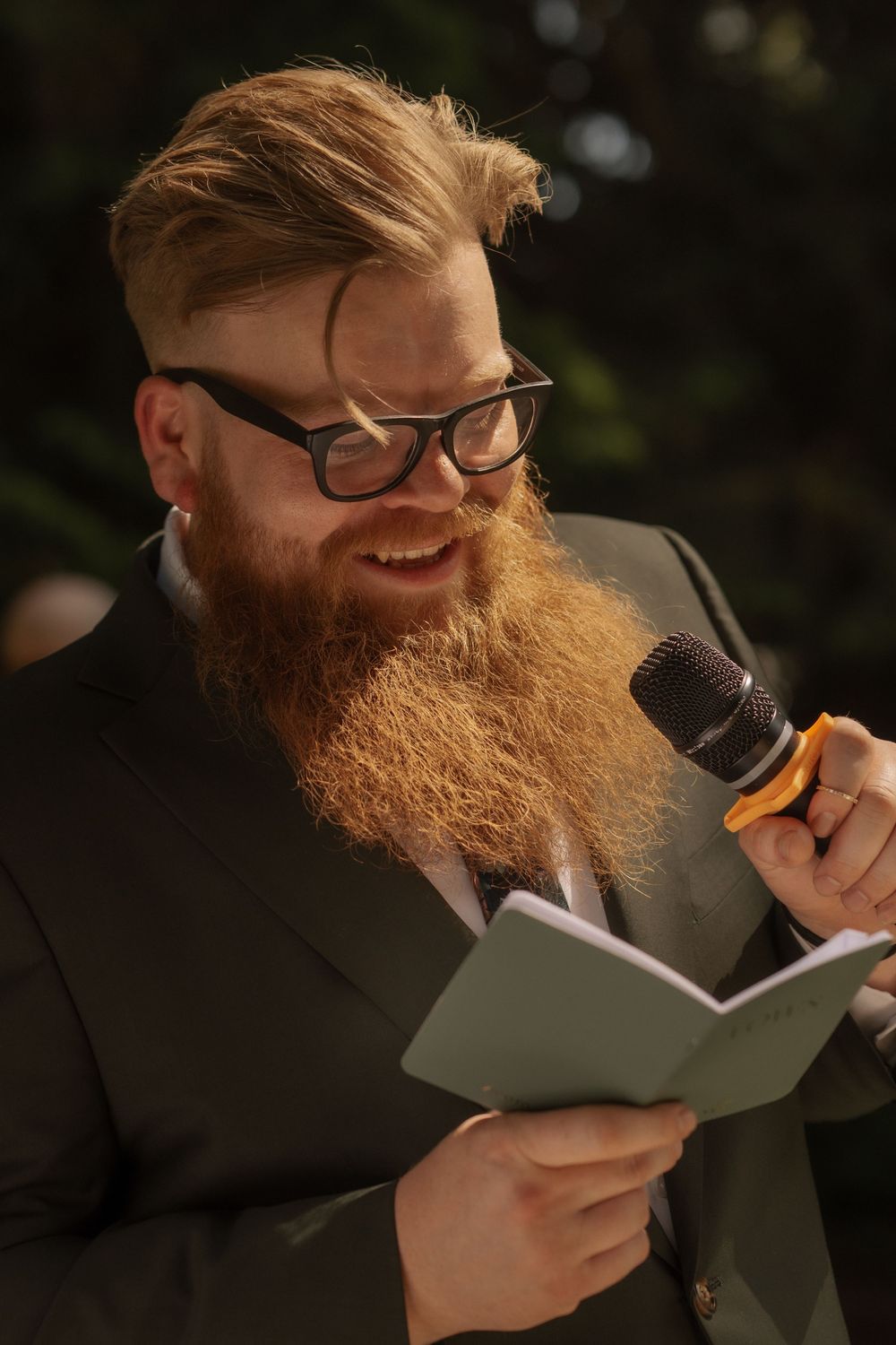 Bearded man in formal wear smiling while reading from a book and holding a microphone outdoors.