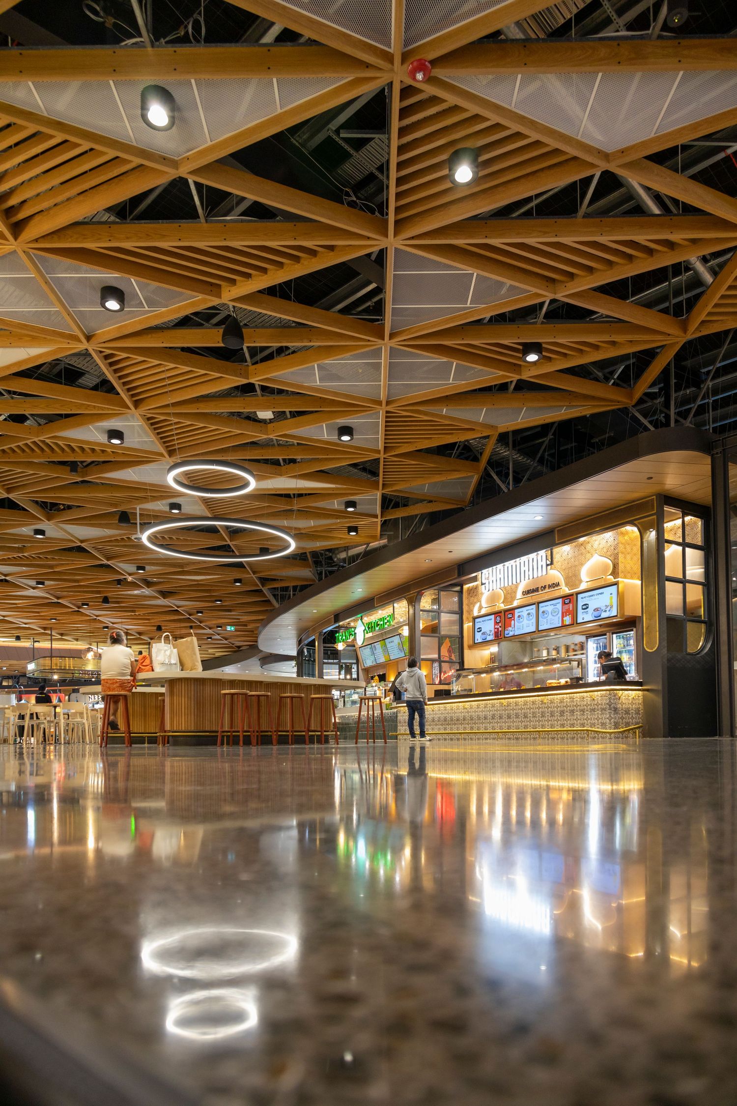 Stylish airport food court with polished floors, wood ceiling lattice, and ring lights reflecting on the ground.