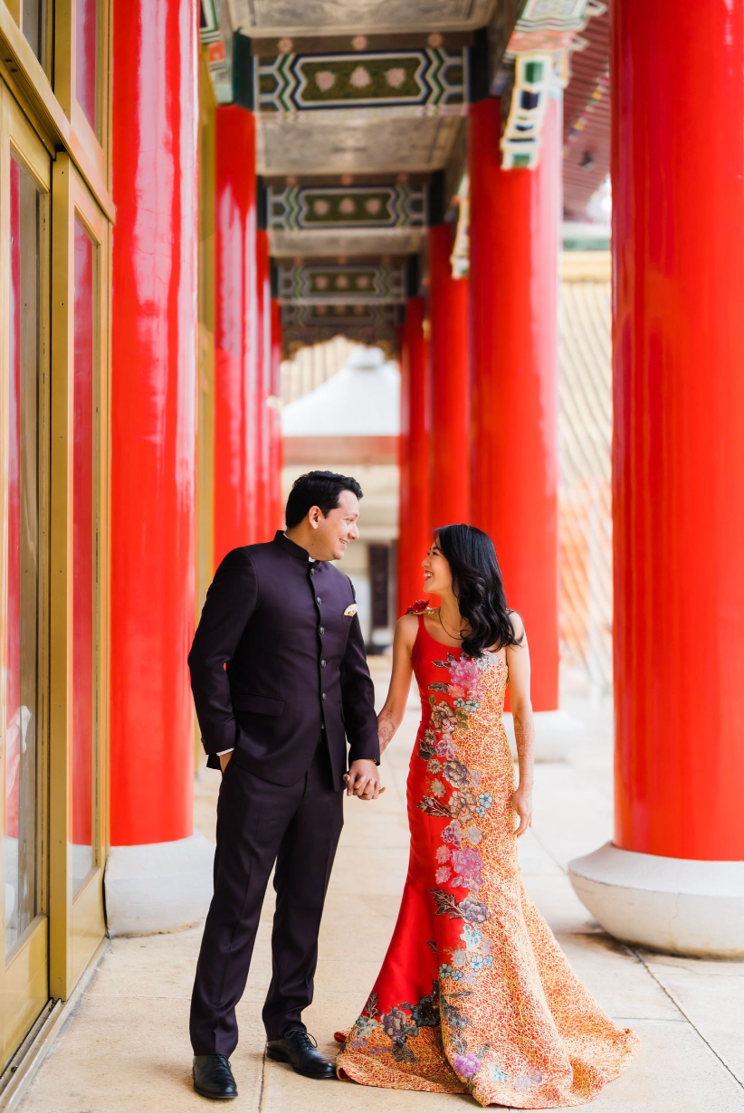 Couple in traditional wedding attire walking under red pillars at an Asian temple pavilion with ornate details.