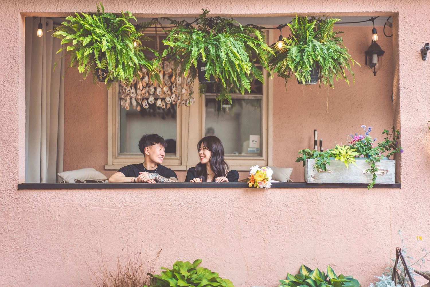 Couple relaxing together at window surrounded by hanging plants and pink walls.