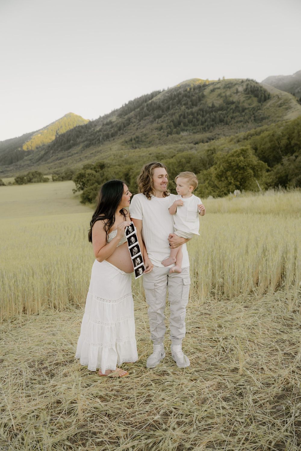An expecting couple shares a joyful moment with their child in a sunlit mountain field.