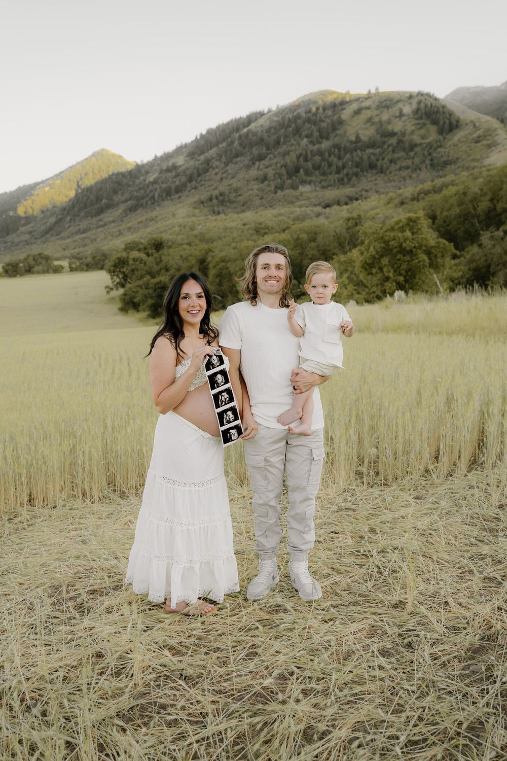 Family of three poses in mountain field with ultrasound strip showing pregnancy announcement.