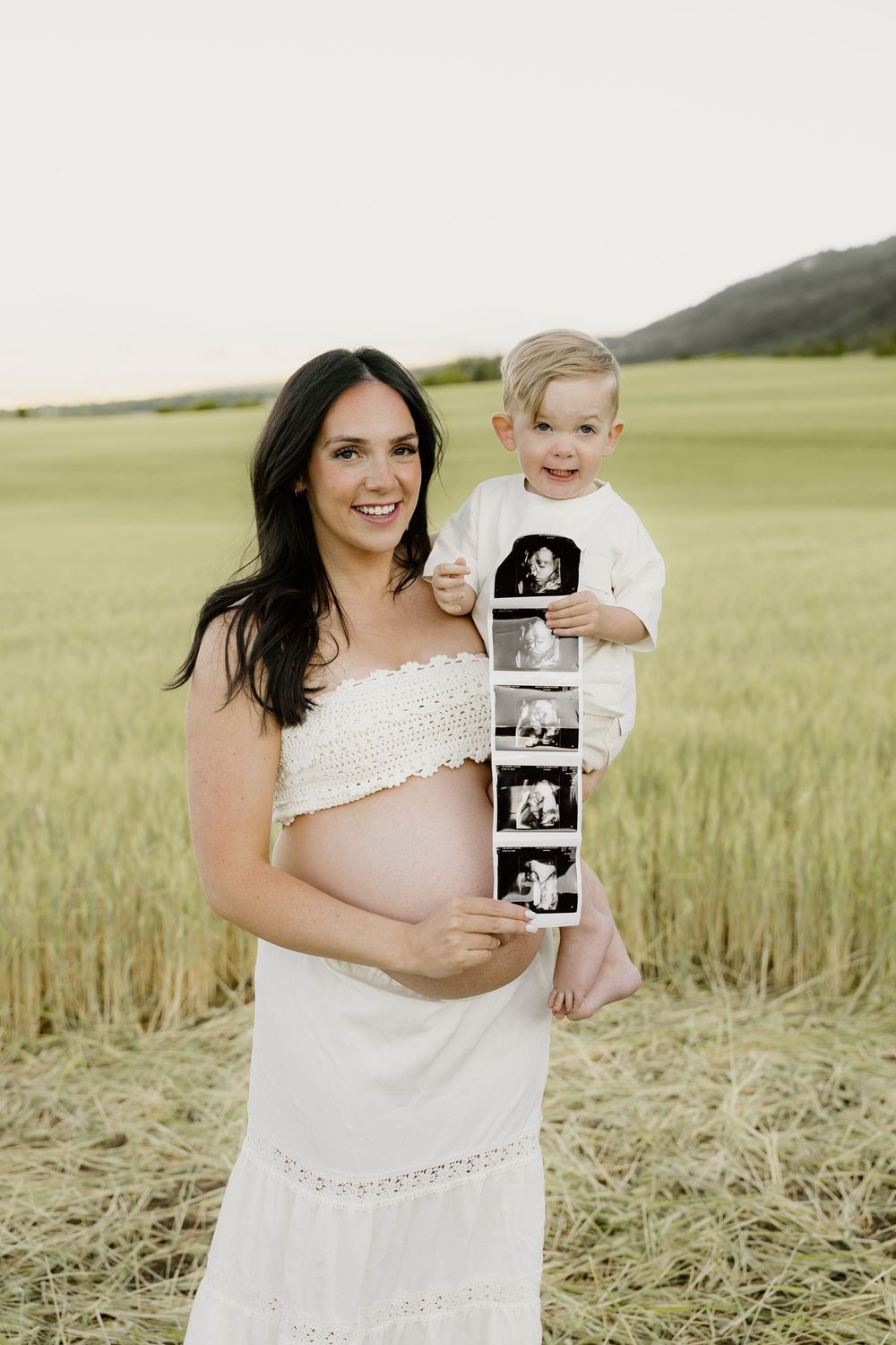 Pregnant woman in white dress holds toddler displaying ultrasound strip in mountain field.