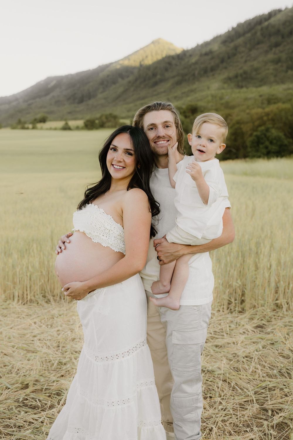 Expectant family poses together in white outfits against a mountainous landscape at sunset.