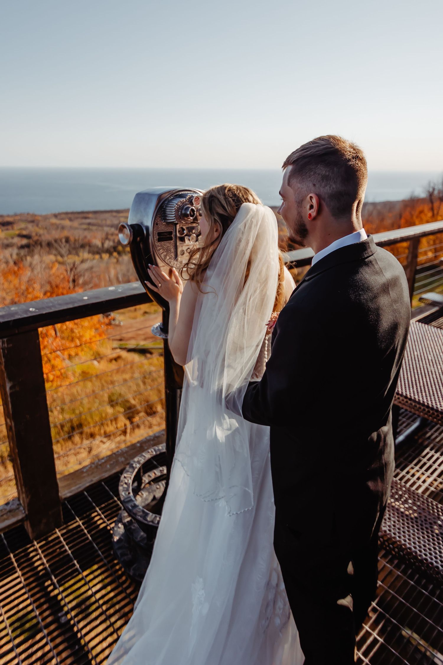 A bride wearing a flowing white veil and groom looking through scenic overlook binoculars during fall sunset.