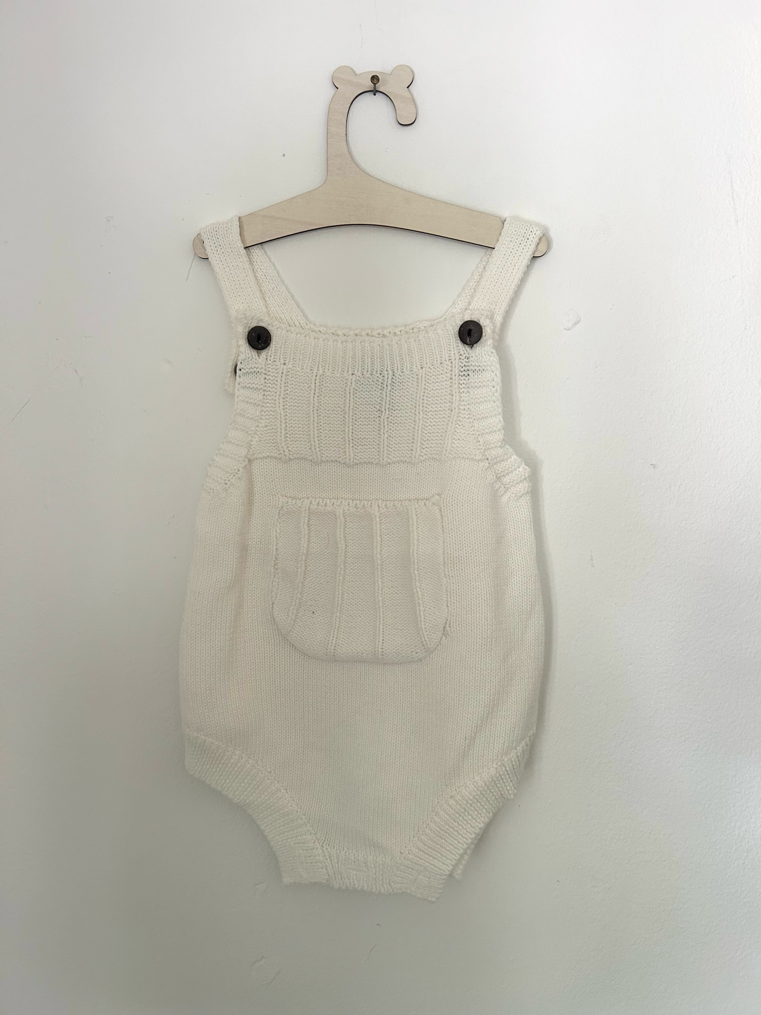 Vintage-style beige knitted baby romper with button details and side pockets hanging on display.