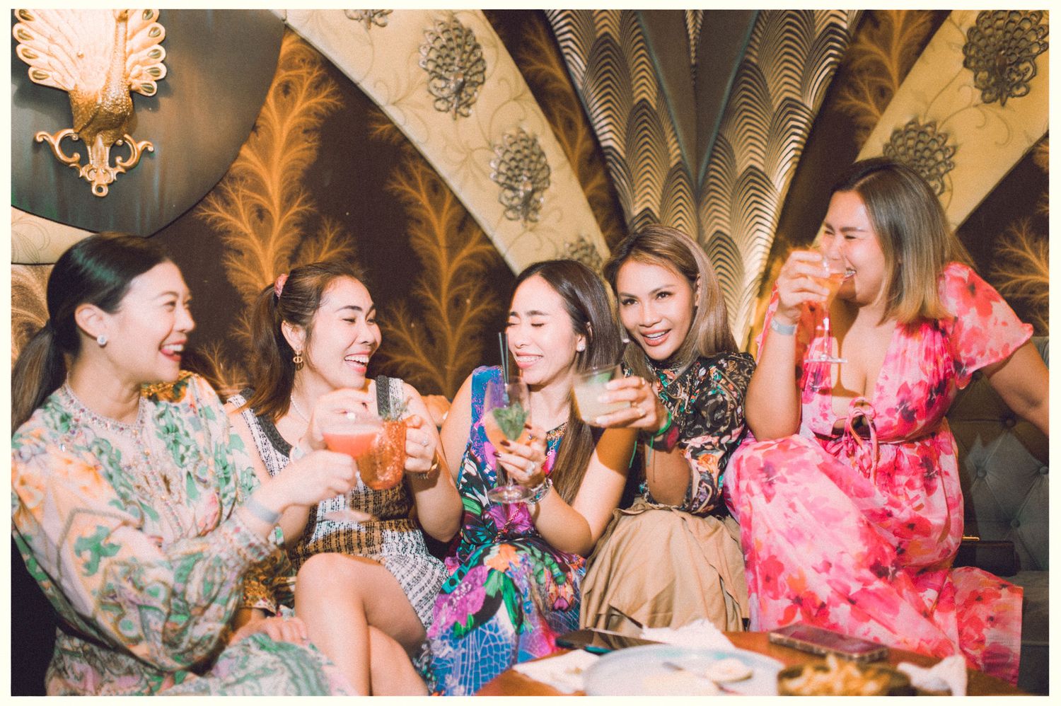 Group enjoying drinks and conversation in floral dresses at social gathering.
