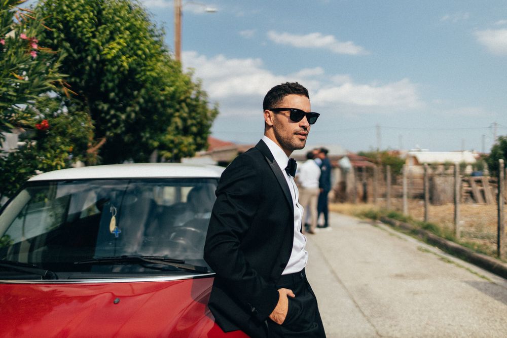 A person in a black suit poses against a red Mini Cooper on a sunny day.