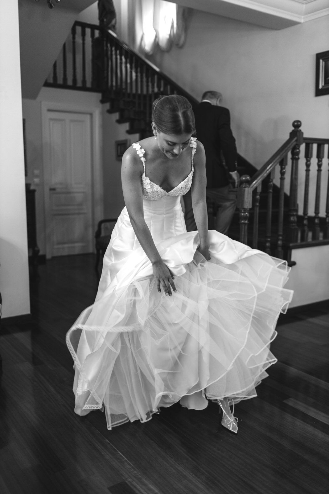 Black and white photo of wedding dress being adjusted on hardwood floor near stairs.
