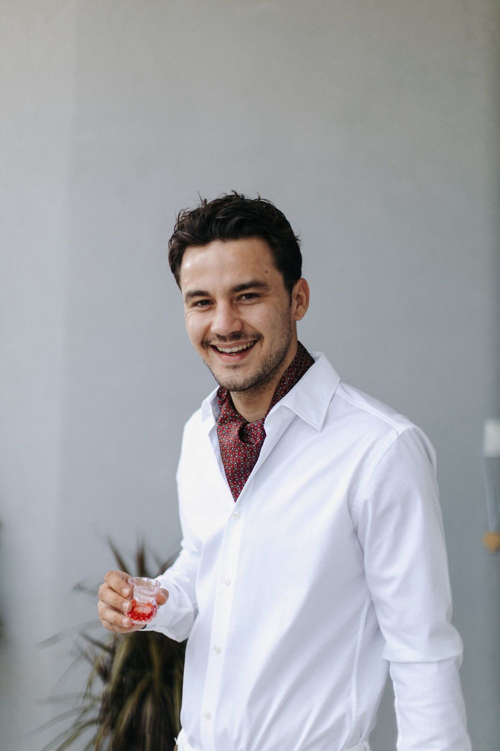 Person wearing a white dress shirt and patterned ascot smiles against neutral background.