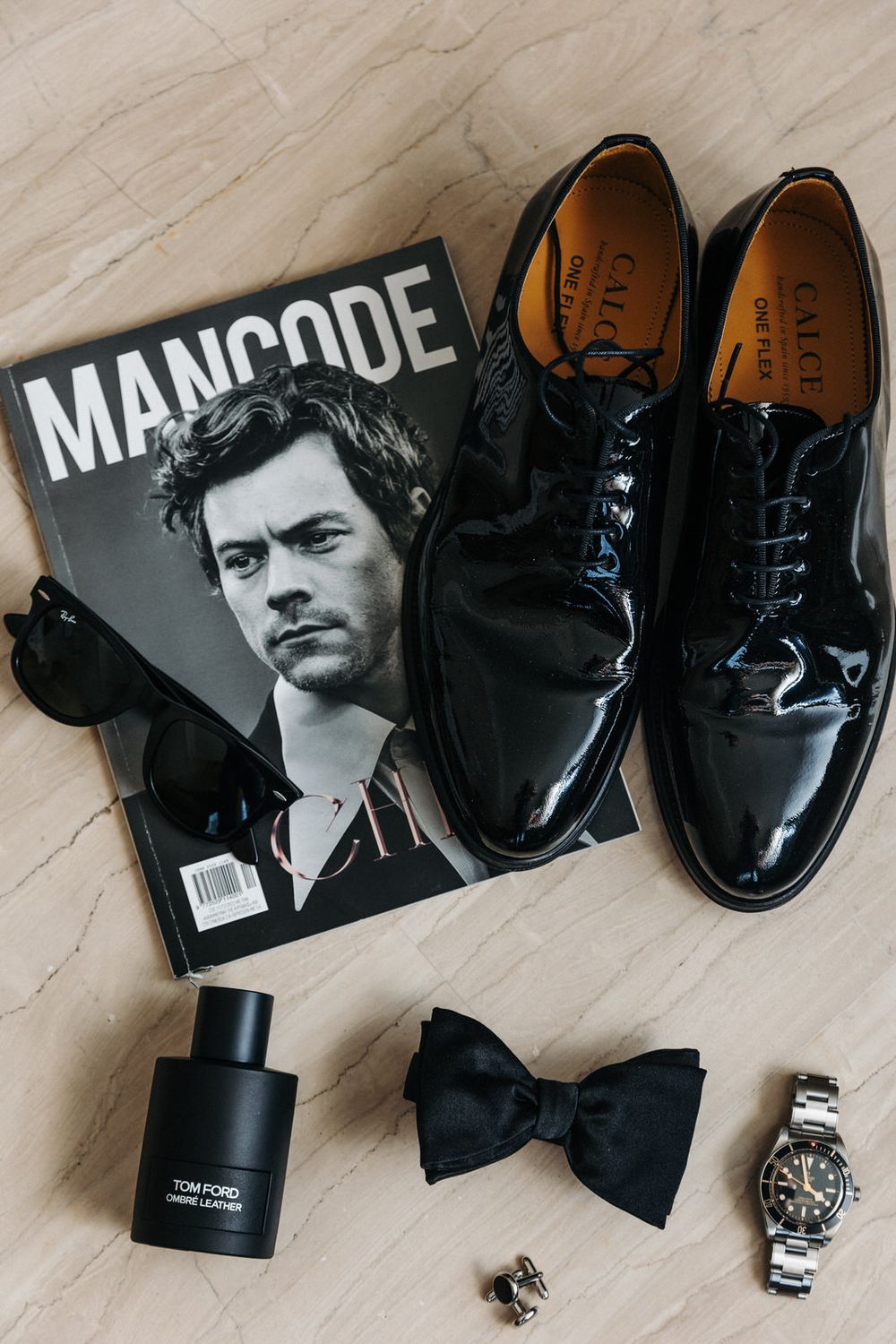 Men's formal accessories including patent leather shoes, bow tie, watch and magazine on marble surface.