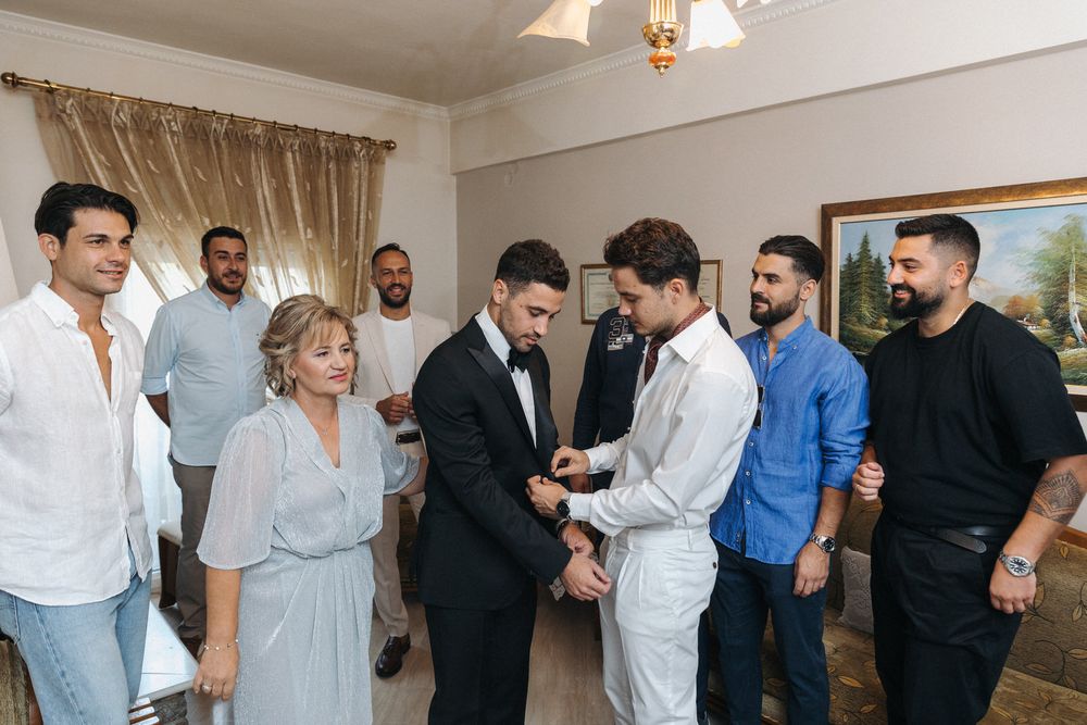 A group gathered for wedding preparations as two people button up formal attire.