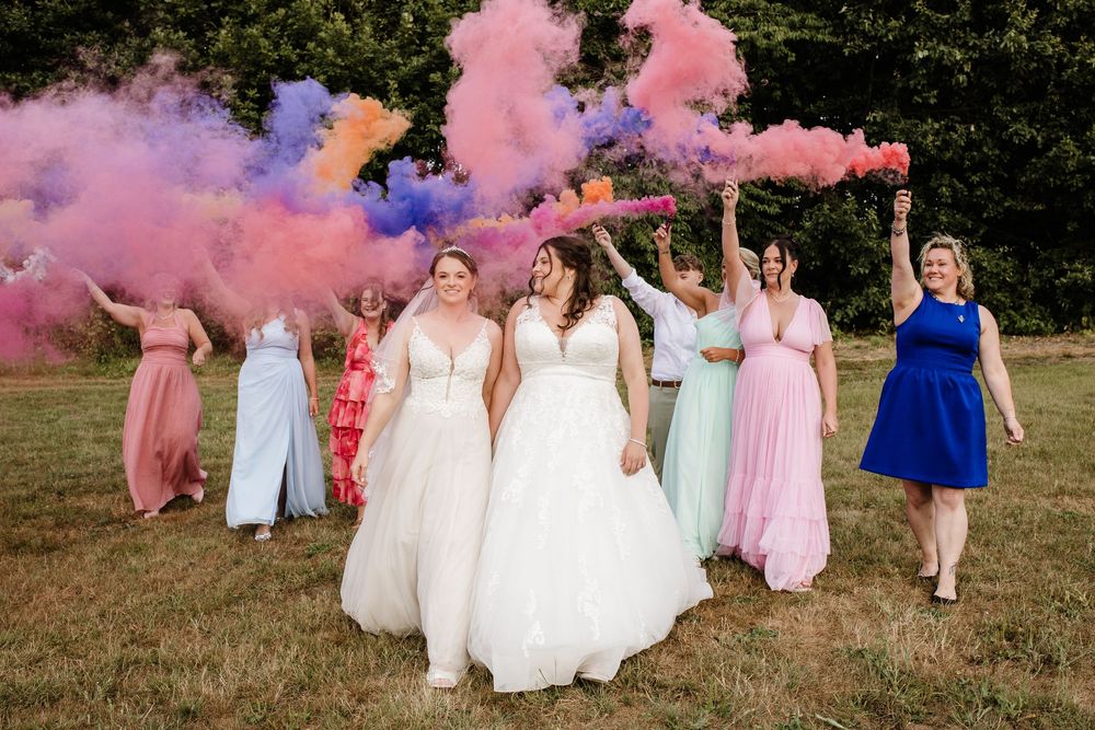Two brides in white gowns walk with colorful smoke bomb bridesmaids in pink, mint, and blue dresses outdoors.