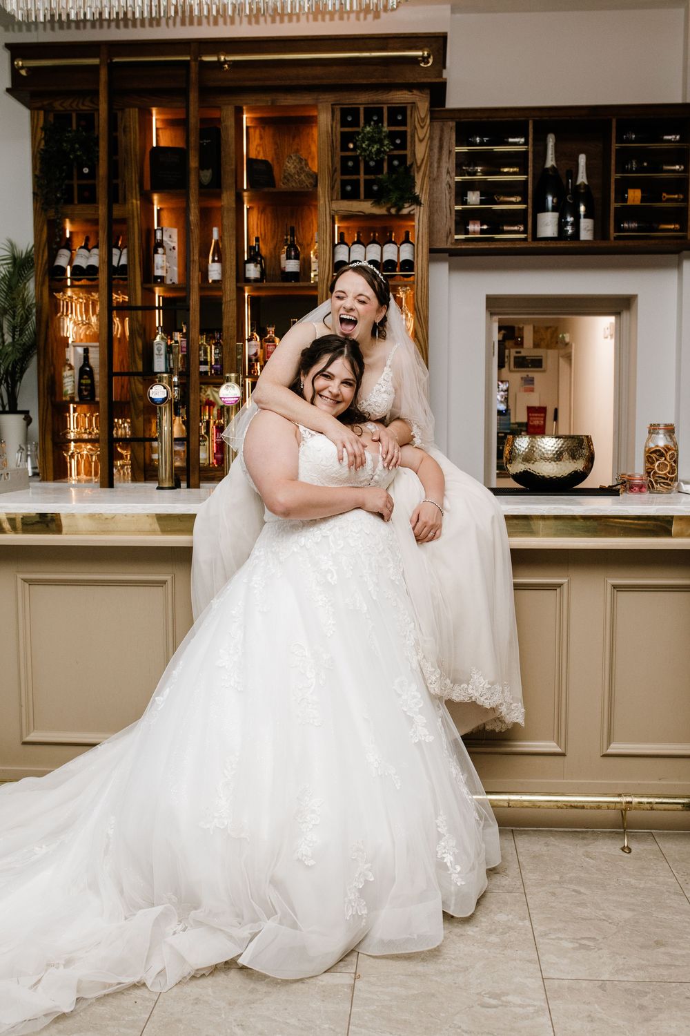Two brides in white wedding gowns laugh joyfully together at a bar, celebrating their wedding day.