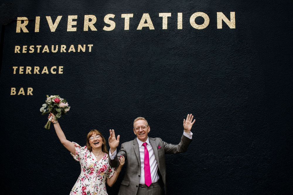 Joyful couple celebrating outside Riverstation Restaurant Terrace Bar, bride holding bouquet, both waving with excitement.