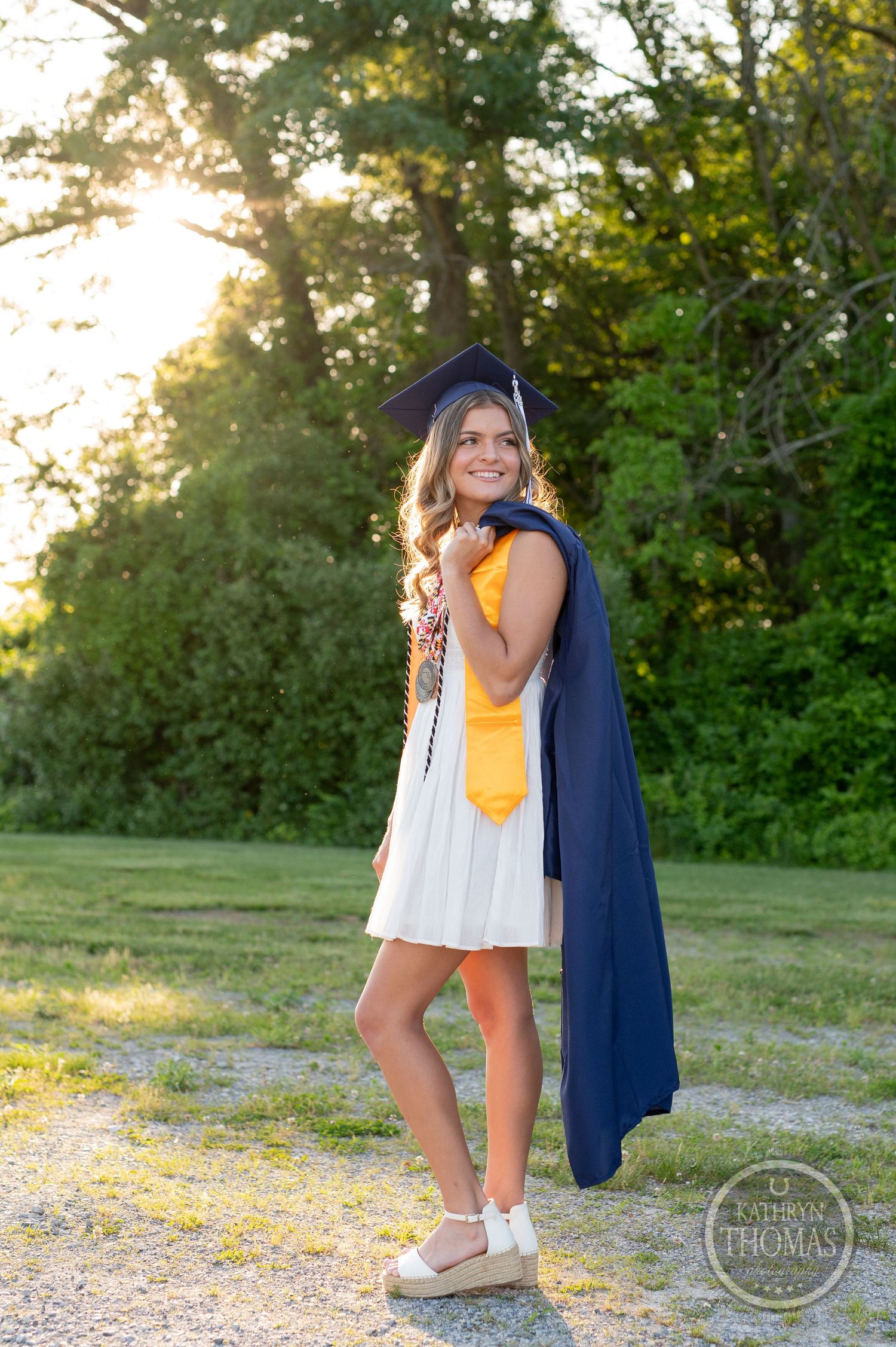 Graduate wearing yellow and white dress with navy cap and gown poses in sunlit garden setting.