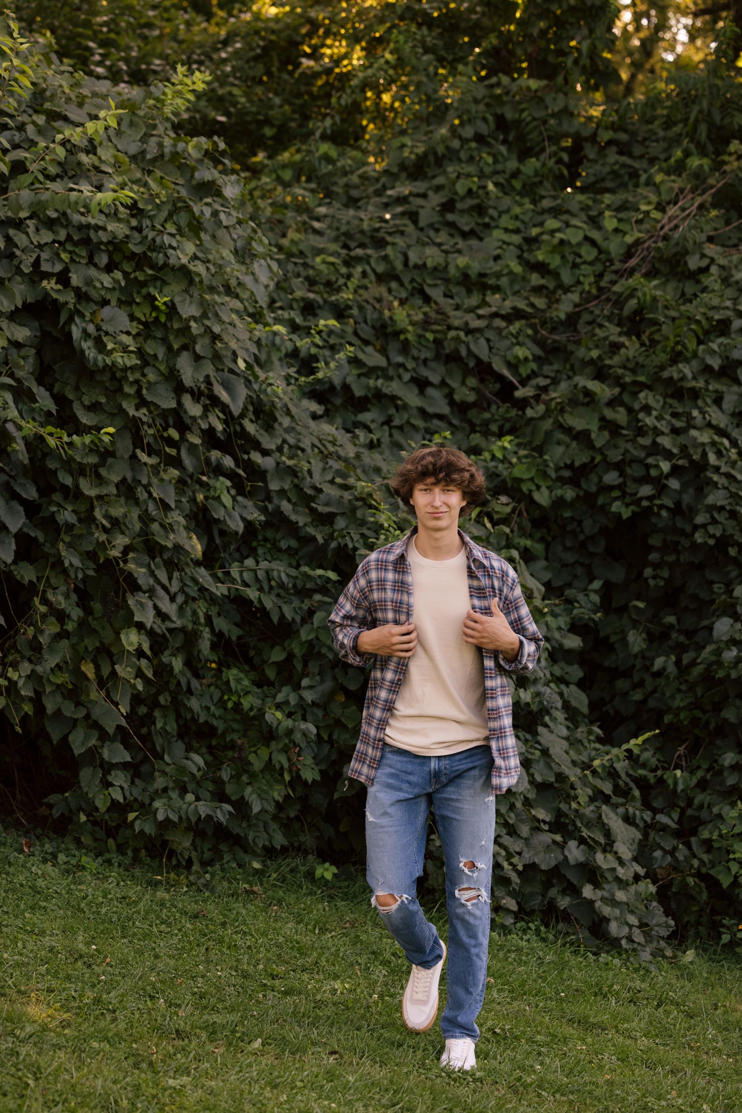 A person in a plaid shirt and jeans walking on grass with leafy foliage in the background.