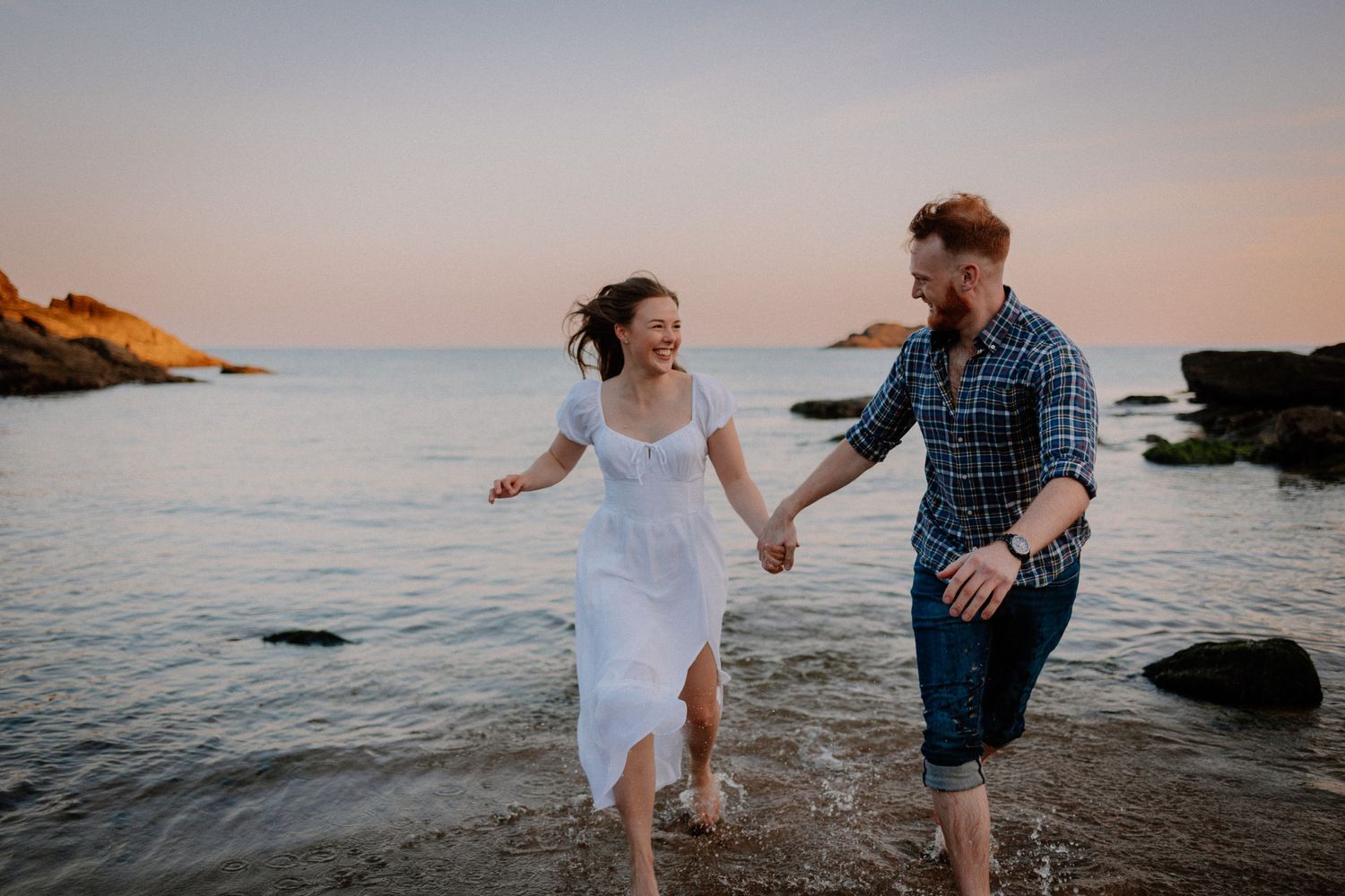 Couple running hand in hand through waves on rocky beach at sunset, Soar Mill Cove, South Hams, Devon