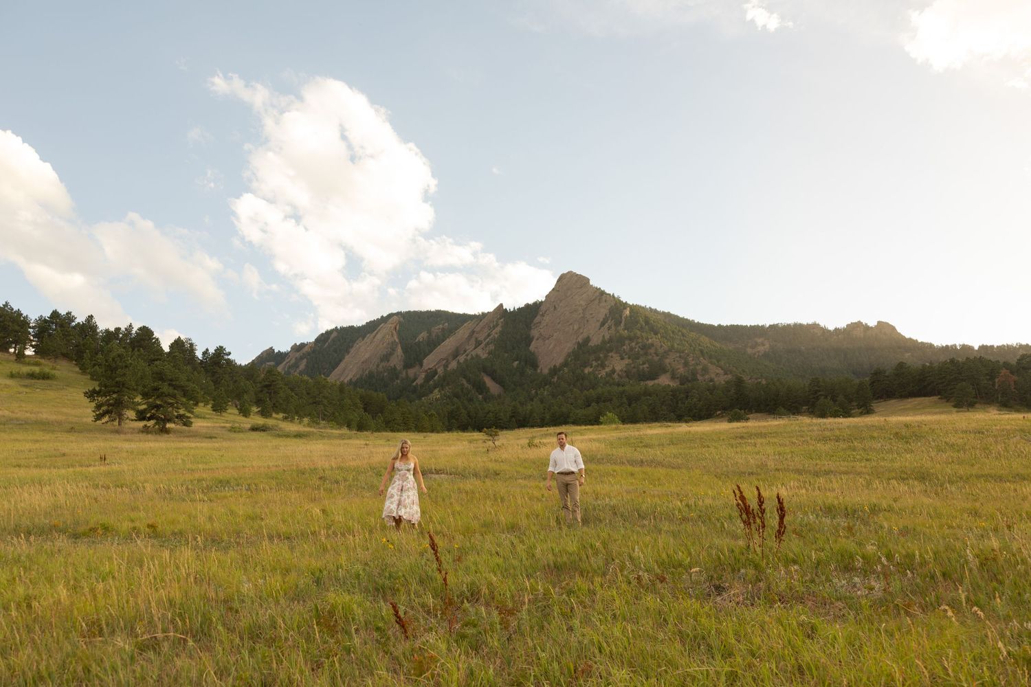 A couple walks through a golden meadow with the iconic Flatirons mountain formation in Boulder rising in the background.