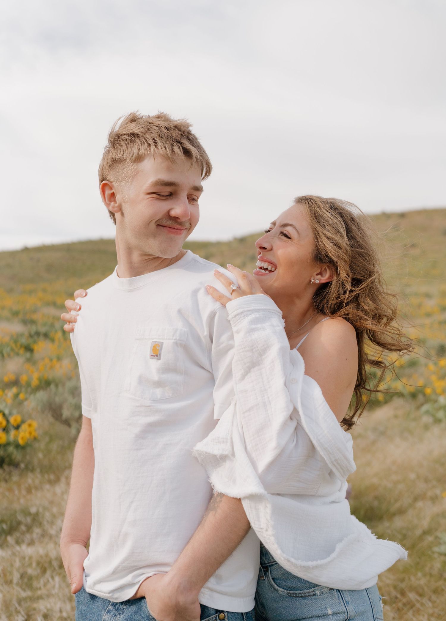 Couple in matching white tops and jeans share playful candid moments in a sunny meadow with wildflowers.