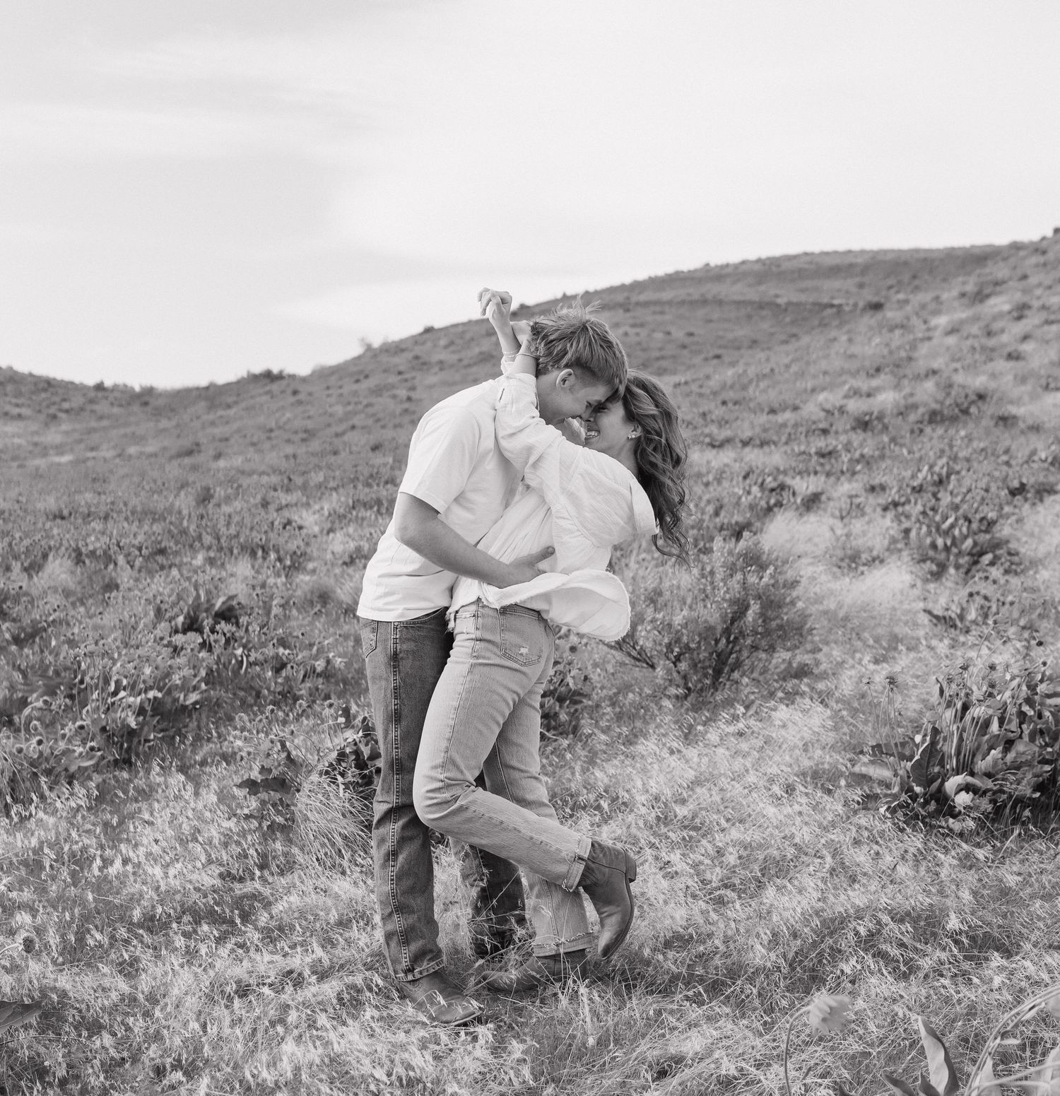 A couple in white clothing embraces and dances together on a grassy hillside in a romantic sequence of movements.