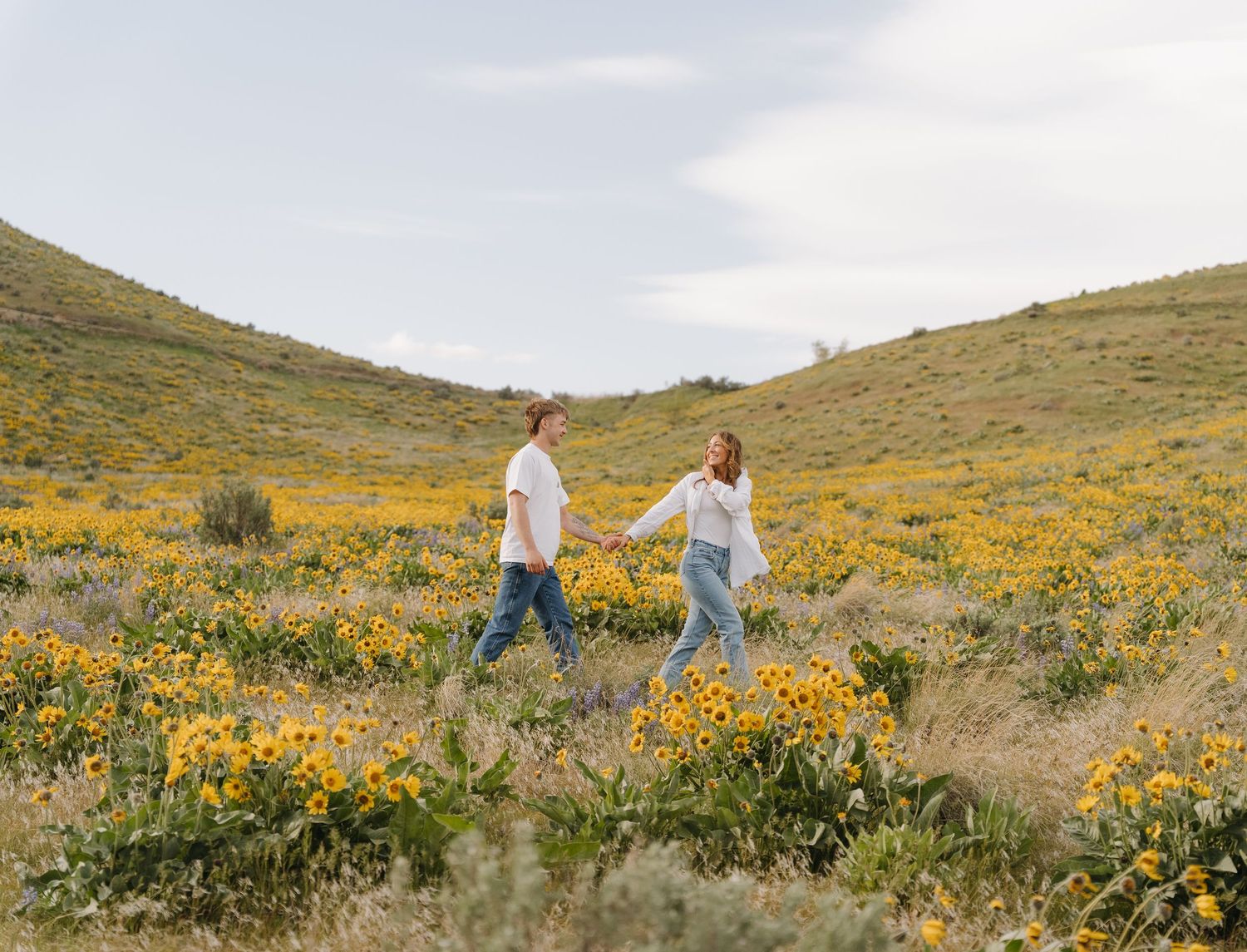 Two people walking through a vibrant hillside meadow filled with blooming yellow wildflowers on a cloudy day.