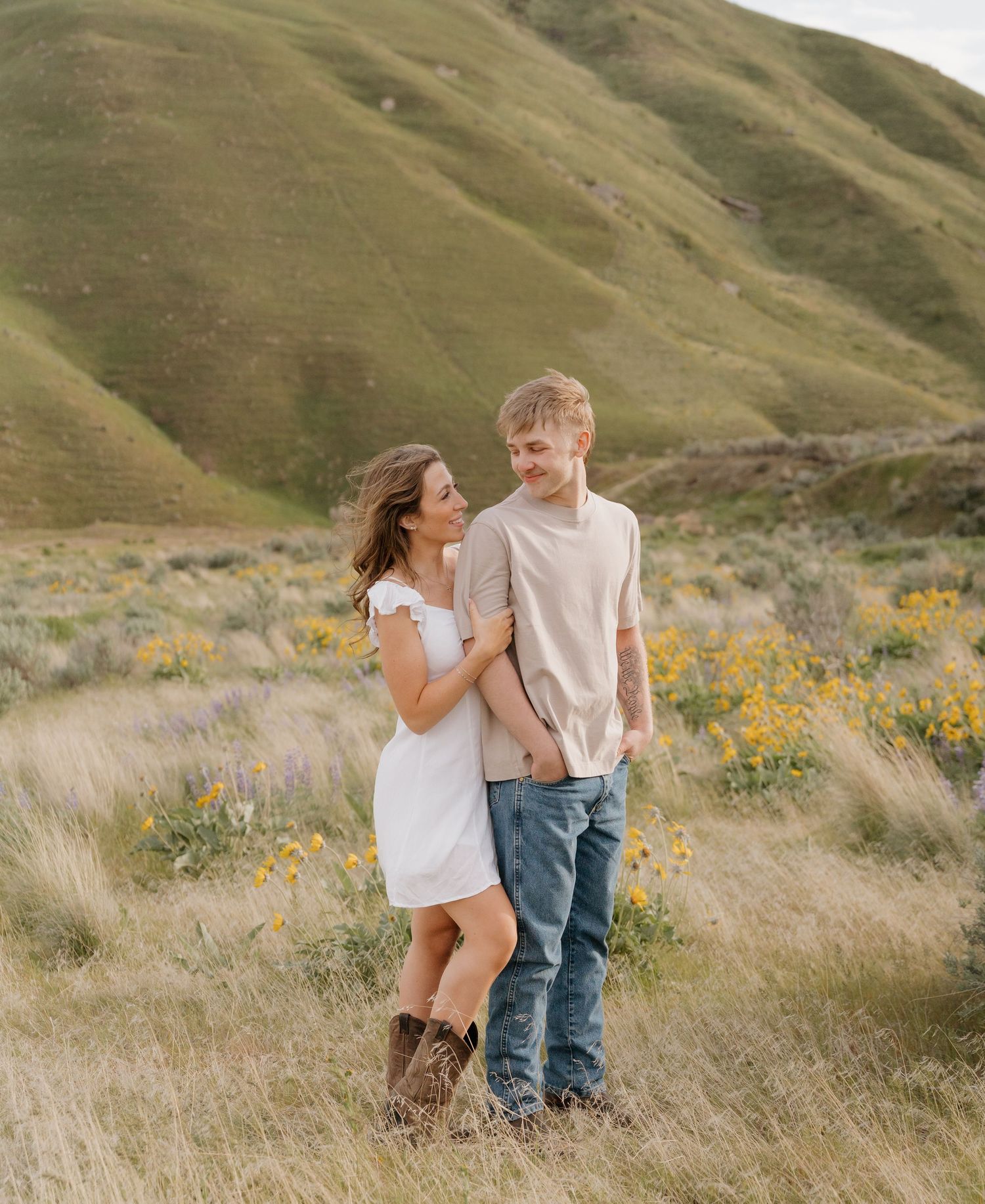 A couple shares intimate moments in a scenic mountain meadow with golden wildflowers and rolling hills in the background.