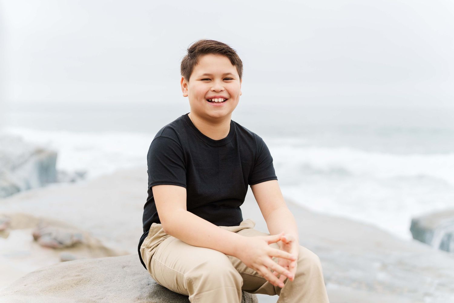 Person in a black t-shirt and khaki pants sits smiling on coastal rocks near the ocean on an overcast day.
