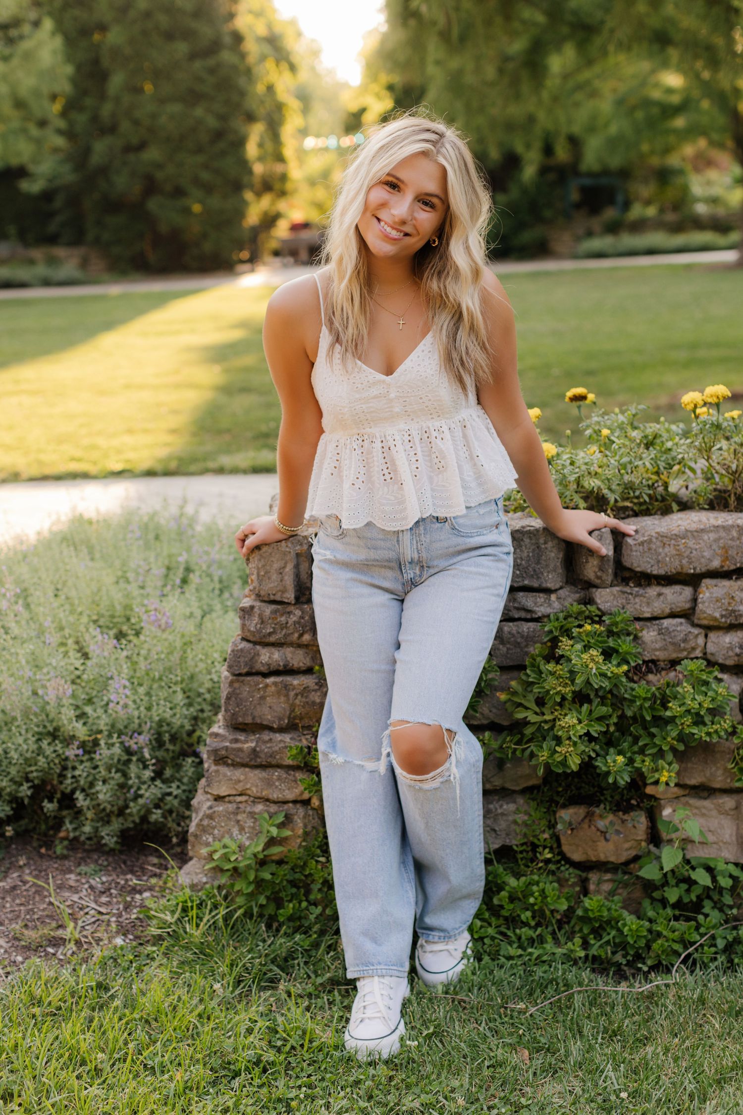 Woman in white peplum top and ripped jeans leans against stone wall in sunny garden.