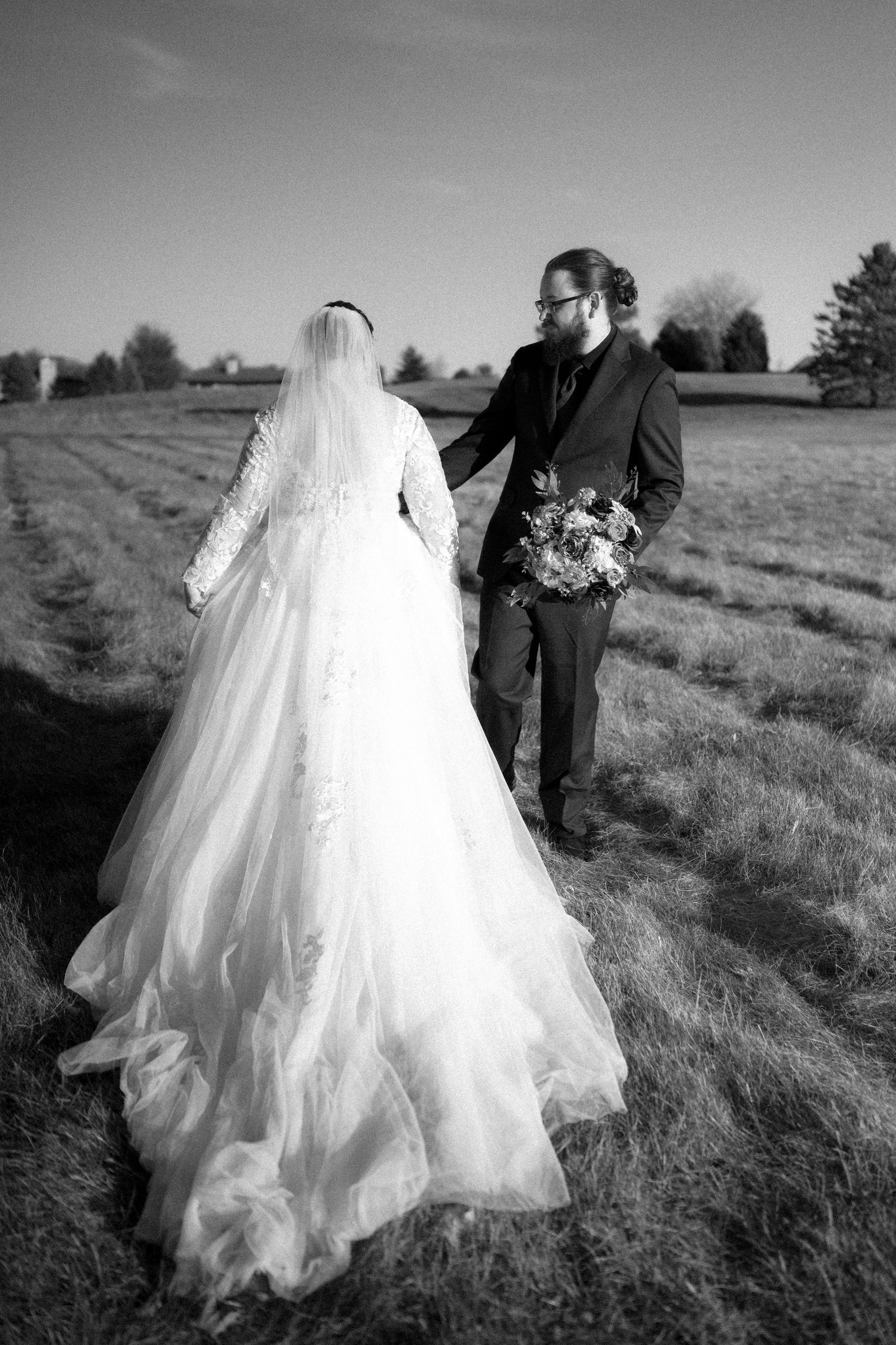 A black and white outdoor wedding photo showing a couple in a field with a stunning lace dress trailing on the ground.