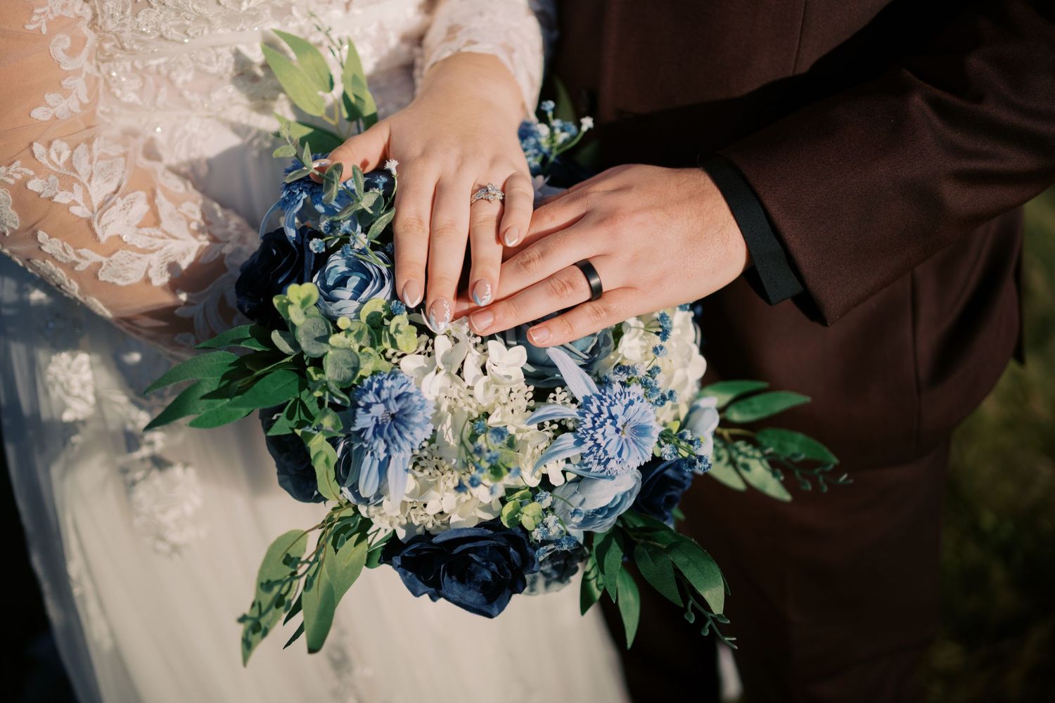 Two hands displaying wedding rings rest on a beautiful bouquet of blue and white hydrangeas with greenery.