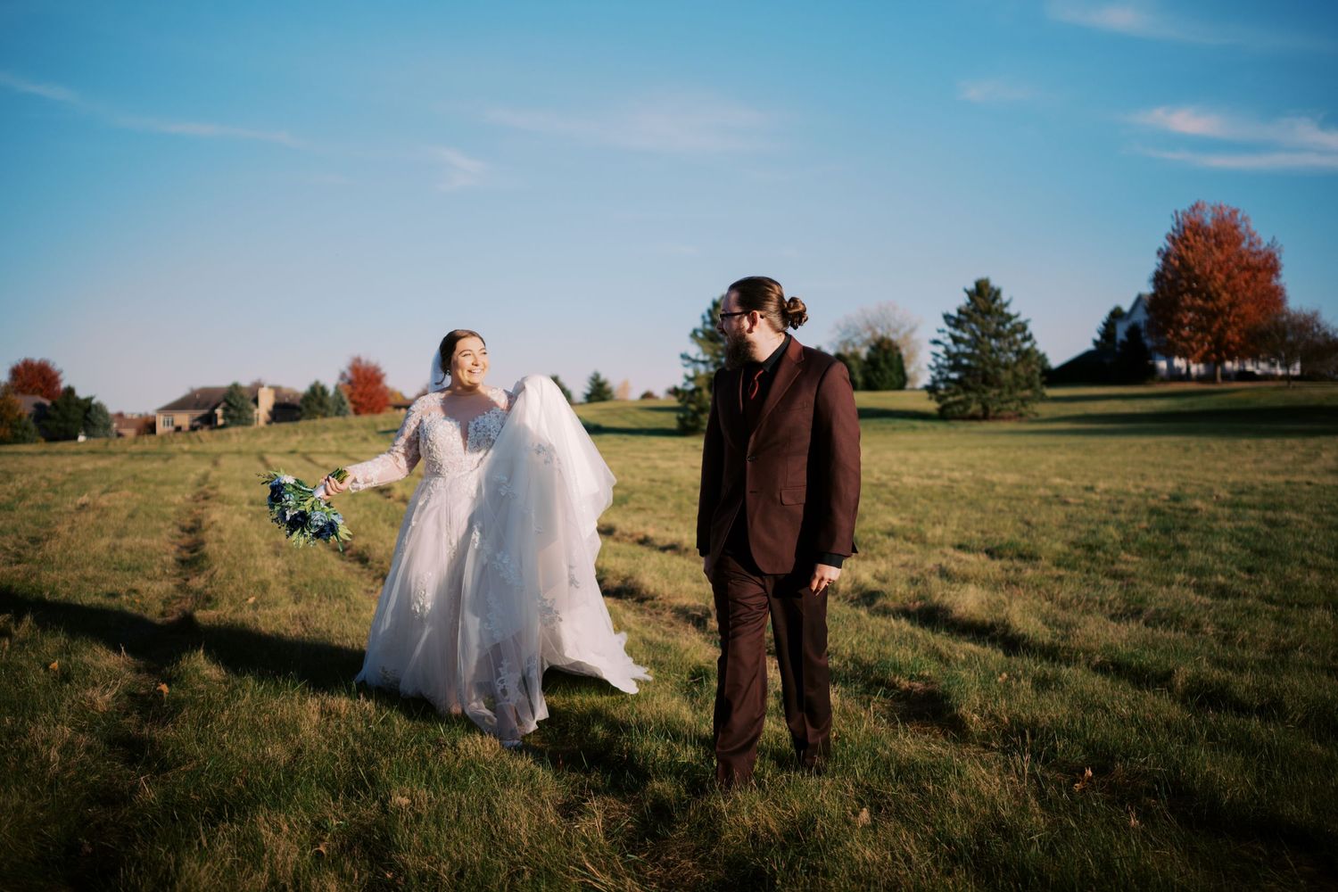 A couple in wedding attire stands in a grassy field on a sunny autumn day with blue skies overhead.